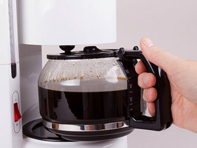 Eine Hand hält eine Kaffeekanne mit frisch gebrühtem Kaffee, die von einer Kaffeemaschine kommt.